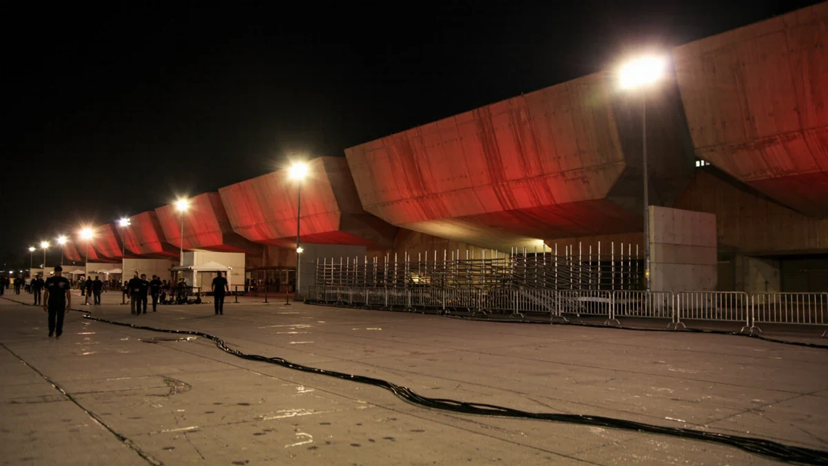 Golden floodlights rake a velvet-roped arrivals carpet as flash bulbs ignite along a wall of marquee glow behind.