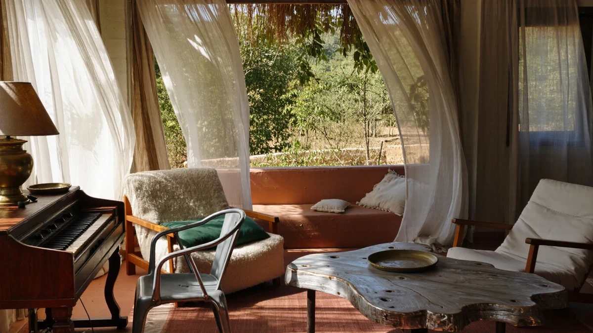 A terrace in low golden light, teak gone soft silver-grey beside a white aluminium sling chair, one person unfolding a linen cover with a watering can at their feet.